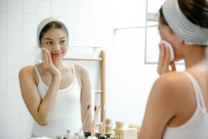 Smiling young Asian female looking at reflection in mirror while cleaning face s