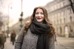 Happy woman in a gray coat and scarf standing outdoors, showcasing a charming sm