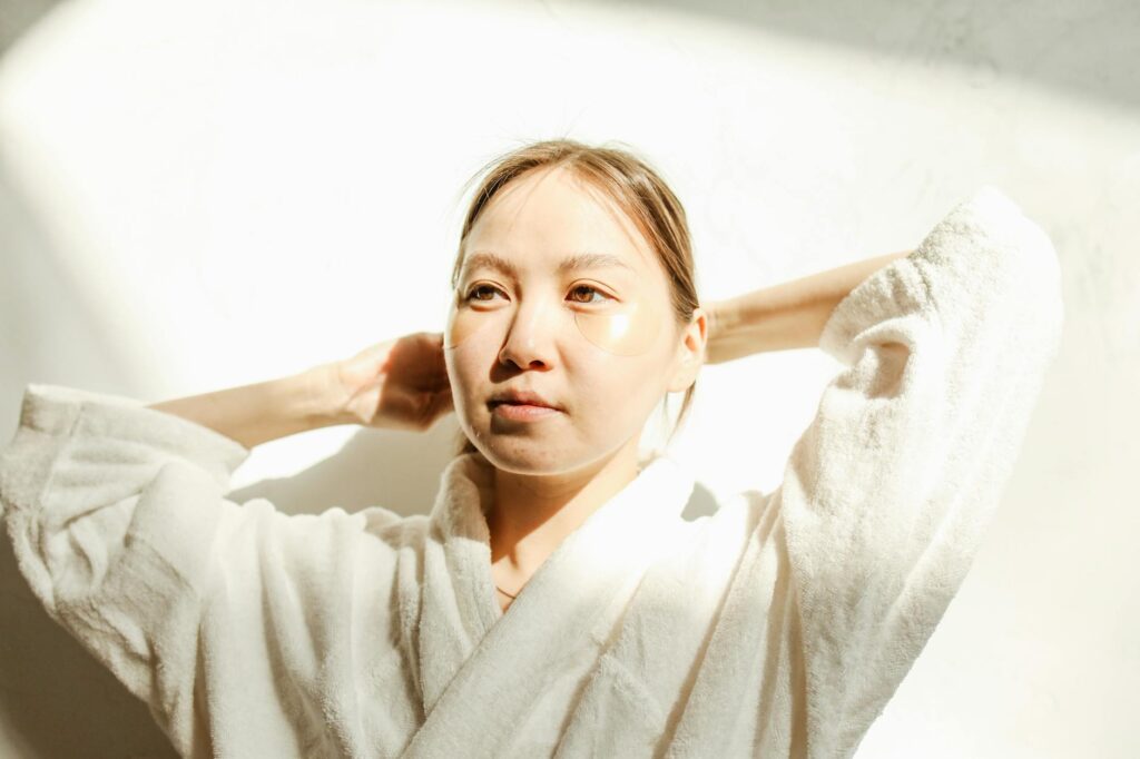 An adult woman in a bathrobe applies under eye masks for skincare routine indoors.