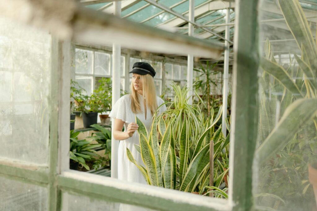 Woman in a white dress and cap exploring a lush greenhouse with various plants.