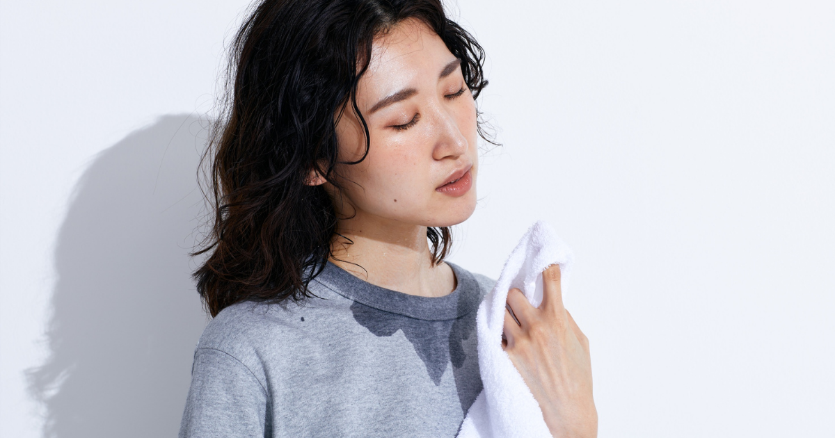 グレーのトップスを着た女性が汗をかきハンカチで顔を拭く・白背景