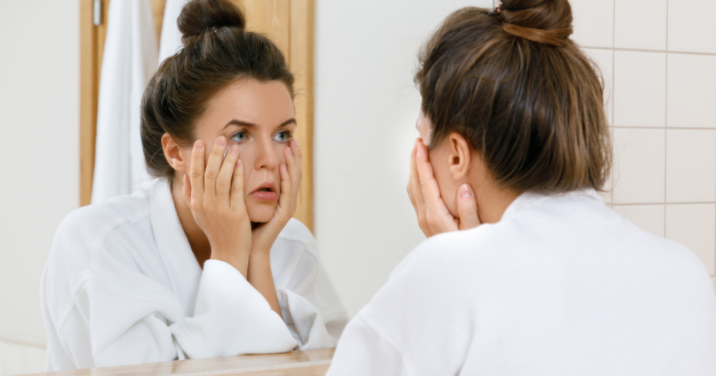 バスローブ姿で鏡の前に両手を顔に当てて肌を確認する女性