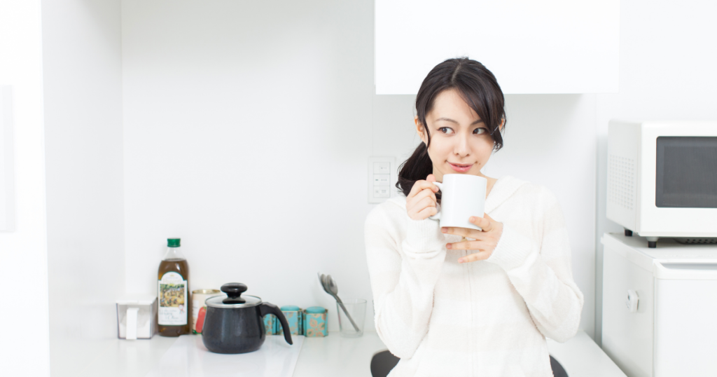 白いセーターを着た日本人女性がキッチンでマグカップを持って微笑む
