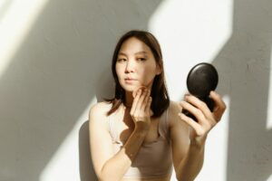 ベージュのタンクトップを着た女性が手鏡でメイクをチェックする・白壁