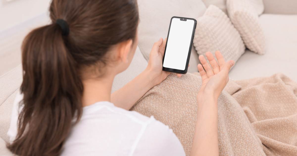 ソファで白紙画面のスマートフォンを持つ後ろ姿の女性 毛布